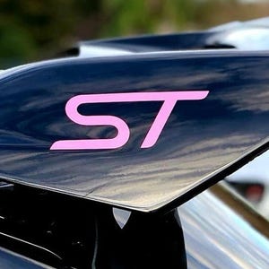 May include: A close-up shot of a black car spoiler featuring the pink letters "ST". The spoiler has a shiny surface, reflecting the sky. The image highlights the spoiler's design and the contrasting pink lettering.