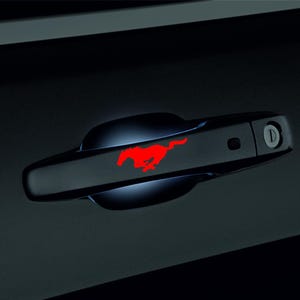 May include: Black car door handle with a red Mustang logo. The handle is sleek and modern, with a keyhole and a small button. The red logo is a striking contrast against the black finish.