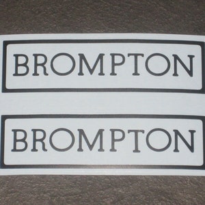 May include: Two white rectangular stickers with the word "BROMPTON" in black capital letters. Each sticker has a black border. The stickers are on a gray surface.