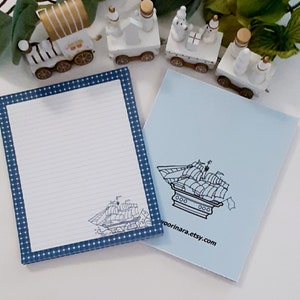May include: A lined notepad with a navy blue polka dot border and a sailboat illustration. A light blue notepad with a sailboat illustration and the text "voorinara.etsy.com" is next to it. Decorative white train figurines are in the background.