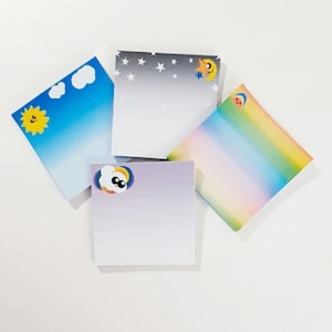 May include: Four colorful sticky note pads with different designs. The first pad has a blue sky with a yellow sun and a white cloud. The second pad has a gray sky with white stars and a yellow moon. The third pad has a rainbow gradient. The fourth pad has a gray gradient with a white cloud and a rainbow.