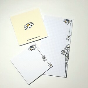 May include: Three white notepads with a black and white daisy and bee design. The notepad on the left has a single daisy, the notepad in the middle has a bee and daisy design, and the notepad on the right has a bee and daisy design along the right edge. The notepad on the right has a grid pattern. The notepad on the left has the text "www.wagrinzara.etsy.com" printed on the bottom.