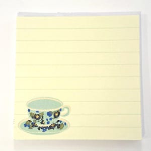 May include: A lined notepad with a decorative teacup and saucer illustration. The teacup and saucer are light blue with a floral pattern in dark blue and green. The notepad has horizontal lines and a light yellow background.