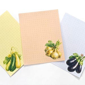 May include: Three rectangular sheets of stationery paper featuring a subtle dot grid pattern. The left sheet is pale yellow with green and yellow squash, the center is peach with yellow onions, and the right is light blue with dark purple eggplants.