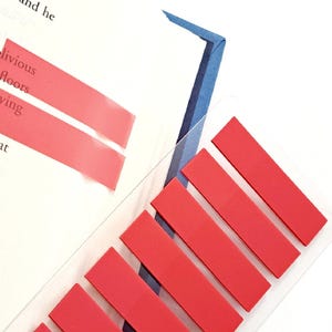 May include: Red sticky notes and a clear bookmark are placed on a book page. The bookmark has a blue and white design. Two red sticky notes highlight text on the page. Additional red sticky notes are attached to the bookmark.