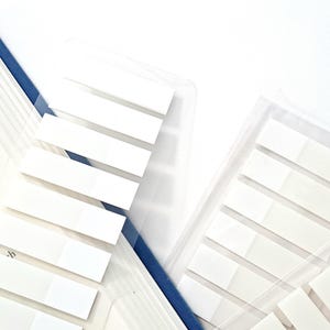 May include: White rectangular sticky page markers are shown next to an open book with a blue spine. The markers are in clear plastic packaging. The book pages are white with black text.