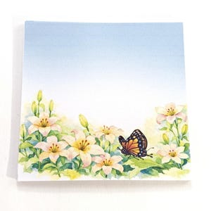 May include: Square notepad with a watercolor illustration of white and yellow lilies, green leaves, and a butterfly with orange, black, and white wings. The background has a gradient of light blue to white.