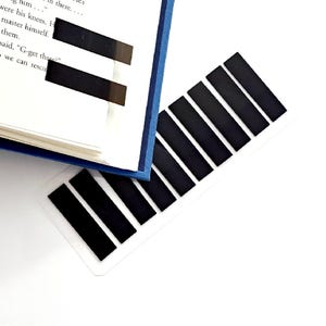 May include: A blue hardcover book with black rectangular page markers. A strip of black markers is next to the book on a white surface. The book is open to a page with text.