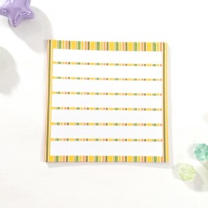 May include: A yellow and green striped notepad with lined paper. The notepad is on a white surface.