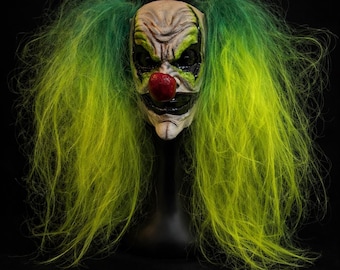 May include: A creepy clown mask with a weathered appearance, featuring a red nose, black accents around the eyes and mouth, and long, neon green and yellow hair. The mask is displayed on a black stand against a black background, creating a spooky effect.