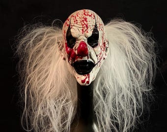 May include: A white and red clown mask with a bloody appearance. The mask has black eyes and a wide, toothy grin. The mask is on a white wig.