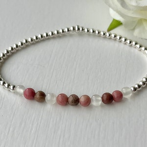May include: A silver beaded bracelet with pink and white gemstone beads. The bracelet is on a white surface.