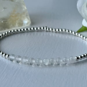 May include: A delicate silver beaded bracelet with clear, faceted beads. The bracelet is made of small, round silver beads and a row of clear, round, faceted beads. The bracelet is on a white surface.