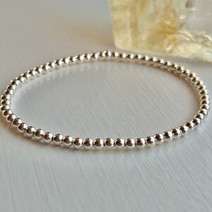 May include: A silver beaded bracelet featuring small, round, shiny silver beads. The beads are strung together on an elastic band, creating a simple and elegant design. The bracelet is displayed on a white surface. The beads are evenly spaced.