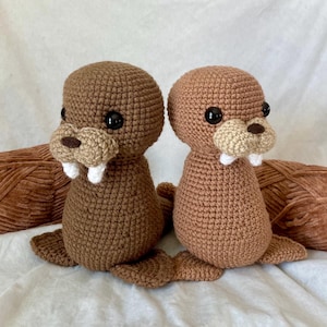 May include: Two handmade crochet walruses in shades of brown. Each walrus has black eyes, a tan snout, and white tusks. The crochet animals are sitting on a white surface with two brown yarn skeins in the background. The walruses are approximately 6 inches tall.