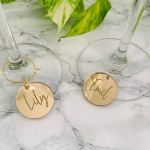 May include: Two circular gold-toned mirrored acrylic wine glass charms featuring custom engraved names. One charm displays the name Lily in a cursive script, while the other displays the name Emil. Each charm is attached to a thin metal hoop.