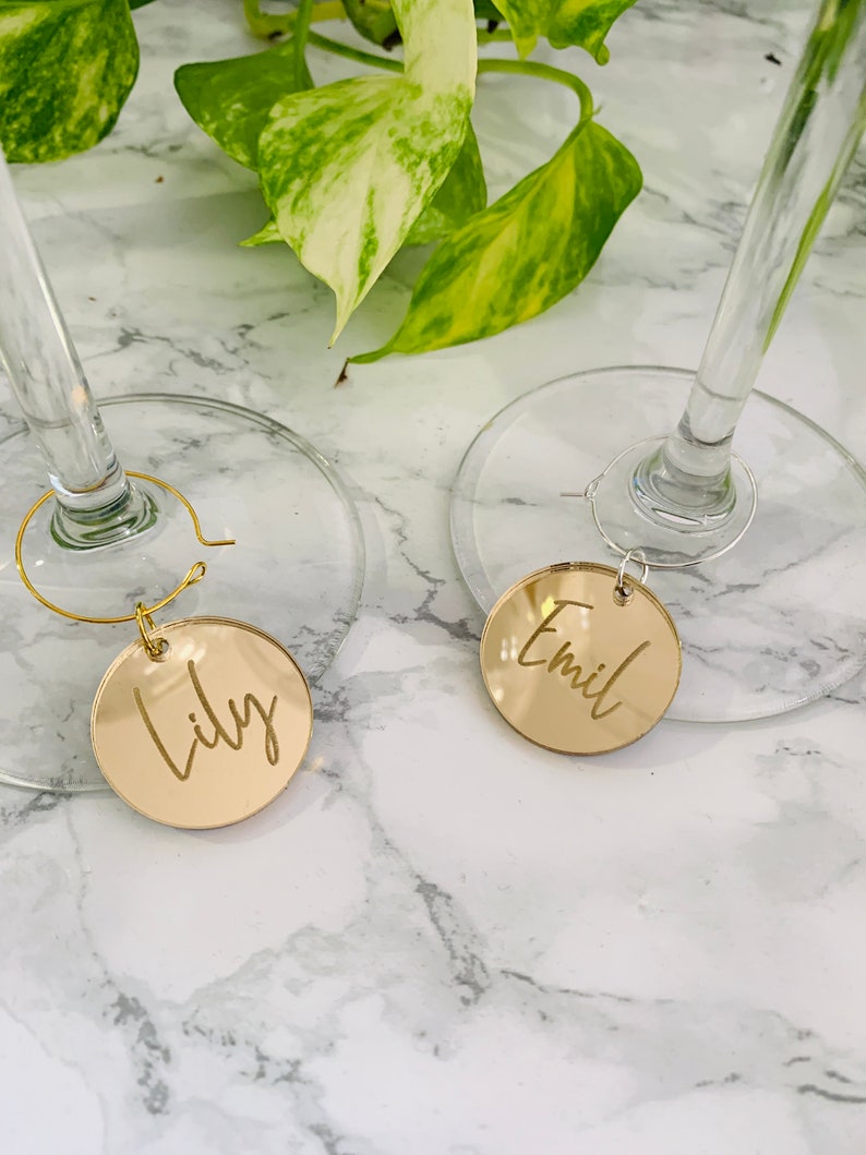 May include: Two circular gold-toned mirrored acrylic wine glass charms featuring custom engraved names. One charm displays the name Lily in a cursive script, while the other displays the name Emil. Each charm is attached to a thin metal hoop.