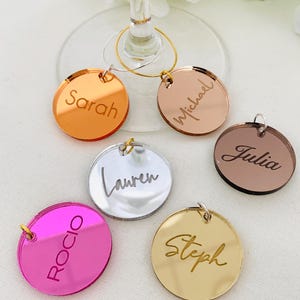 May include: A collection of six circular acrylic wine glass charms in various metallic and vibrant colours, including gold, silver, rose gold, copper, and hot pink. Each charm is engraved with a unique name: Sarah, Michael, Julia, Lauren, Rocio, and Steph.
