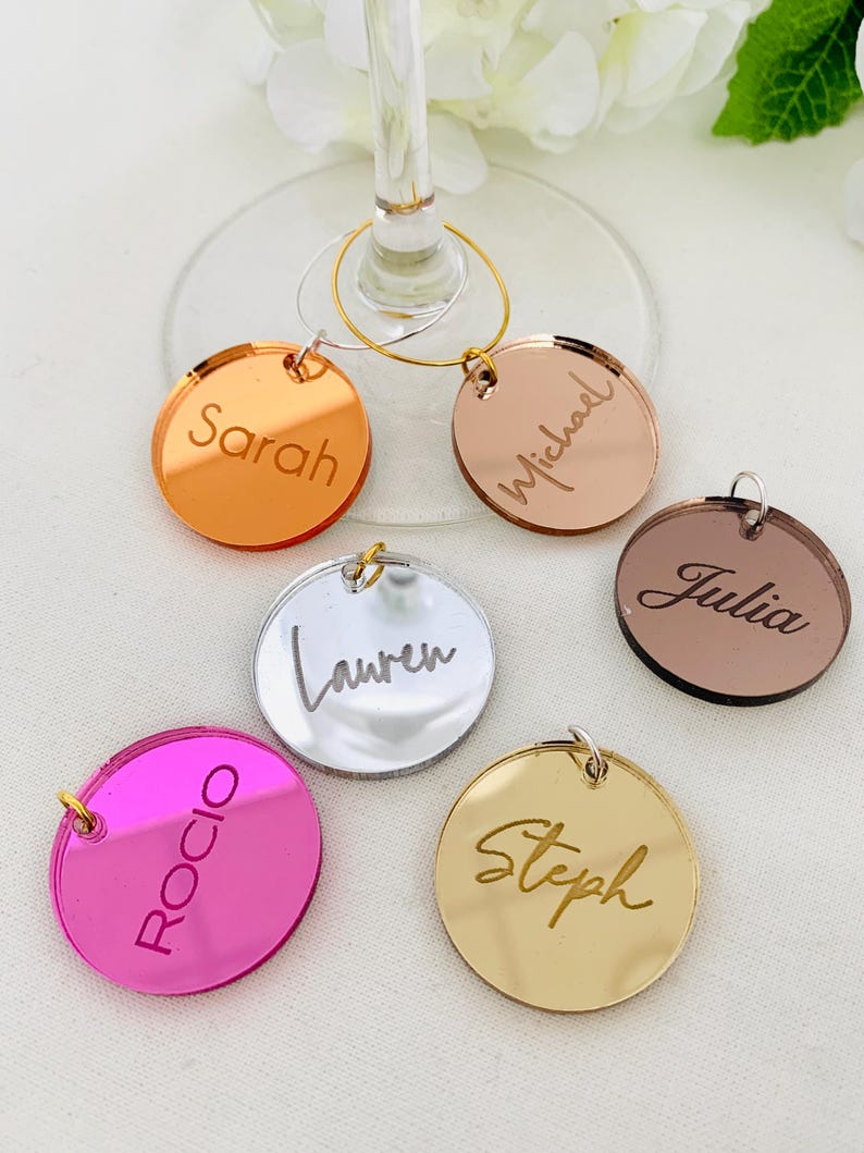 May include: A collection of six circular acrylic wine glass charms in various metallic and vibrant colours, including gold, silver, rose gold, copper, and hot pink. Each charm is engraved with a unique name: Sarah, Michael, Julia, Lauren, Rocio, and Steph.