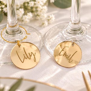 May include: Two circular gold-toned mirrored wine glass charms attached to the stems of two clear glass flutes. Each charm features a custom name engraved in a cursive script, with one charm reading Lily and the other charm reading Emil.