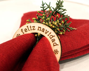 May include: A wooden napkin ring with the text "Feliz Navidad" and a snowflake design. The ring is holding a red napkin and a sprig of green foliage.