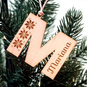 Puede incluir: Adorno de Navidad de madera con forma de la letra M con el nombre "Mariana" grabado en él. El adorno está decorado con diseños de copos de nieve.