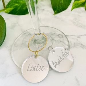 May include: Two circular mirrored acrylic wine glass charms featuring engraved names. One charm is attached to a gold-toned metal hoop and displays the name Louise, while the other is on a silver-toned metal hoop and displays the name Brandon.
