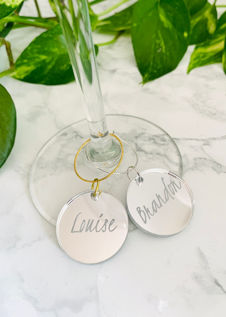 May include: Two circular mirrored acrylic wine glass charms featuring engraved names. One charm is attached to a gold-toned metal hoop and displays the name Louise, while the other is on a silver-toned metal hoop and displays the name Brandon.
