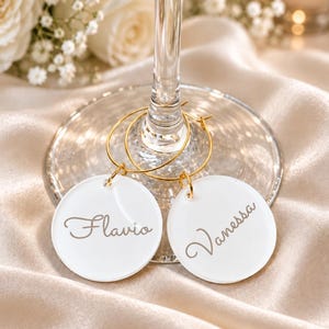 May include: Two circular white acrylic wine glass charms with gold-toned metal hoops. Each charm features a name engraved in a cursive script font, with one charm displaying the name Flavio and the other charm displaying the name Vanessa.