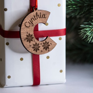 Puede incluir: Un adorno navideño de madera con forma de la letra C con el nombre "Cynthia" grabado en él. El adorno está decorado con copos de nieve y está atado con una cinta roja.