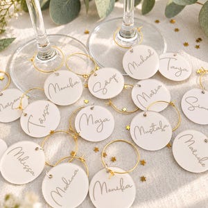 May include: A collection of round white acrylic wine glass charms featuring gold metal hoops and personalised names written in a cursive script. Each charm is circular with a smooth finish, and the set includes various names such as Maja, Melissa, and Medina, accented by small gold star-shaped confetti scattered among the charms.