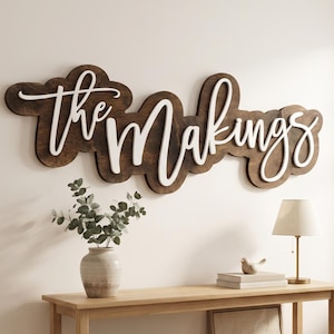 May include: A wall-mounted wooden sign featuring the text "the Makings" in white script lettering layered over a dark brown, cloud-shaped wooden backing. The sign is displayed above a light-colored wooden console table with a shelf.