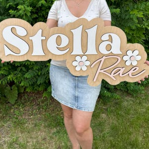 May include: A custom wooden wall sign featuring the name Stella Rae in layered lettering. The word Stella is in white, and the word Rae is in light pink. The sign includes two white and pink flower accents on a light wood-toned backing.