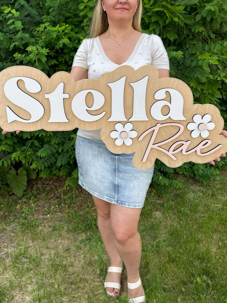 May include: A custom wooden wall sign featuring the name Stella Rae in layered lettering. The word Stella is in white, and the word Rae is in light pink. The sign includes two white and pink flower accents on a light wood-toned backing.