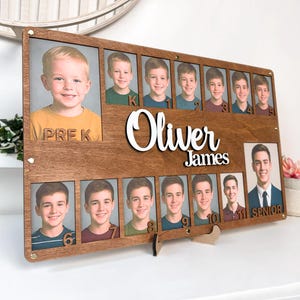 May include: A rectangular wooden school photo display board featuring the name Oliver James in white script. It includes a large slot for a Pre-K photo, eleven smaller slots numbered 1 through 11, and a large slot for a Senior photo.
