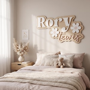 May include: A custom wooden wall sign featuring the name Rory Lewis in layered white and light wood lettering, accented with two white and light pink flower cutouts. The sign is mounted on a wall above a bed with light pink bedding and a teddy bear.