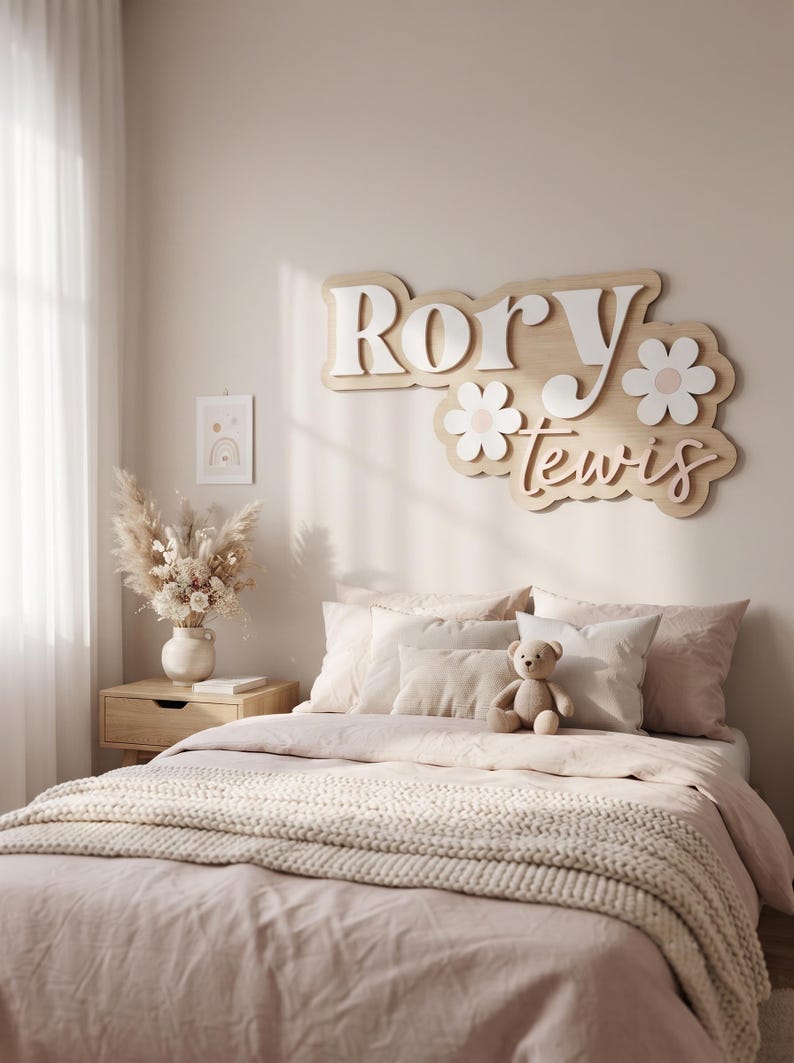 May include: A custom wooden wall sign featuring the name Rory Lewis in layered white and light wood lettering, accented with two white and light pink flower cutouts. The sign is mounted on a wall above a bed with light pink bedding and a teddy bear.