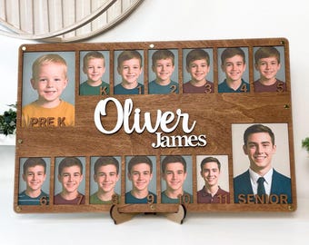 Pode incluir: Um expositor de fotos de madeira com o nome "Oliver James" em escrita branca. Apresenta uma série de fotos escolares do pré-escolar ao último ano, mostrando o crescimento de uma pessoa ao longo do tempo. O expositor é suportado por um suporte de madeira.