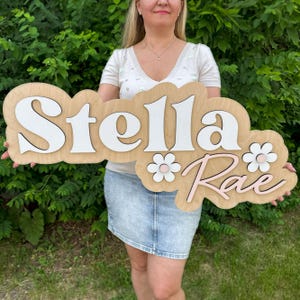 May include: A custom wooden wall sign featuring the name Stella Rae in layered 3D lettering. The word Stella is in white, while Rae is in light pink. The sign includes two white flower accents with pink centers, all mounted on a natural wood base.
