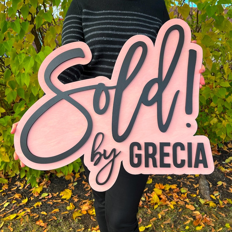 Op de afbeelding: Een groot, lichtroze, op maat gevormd houten bord met de zwarte, verhoogde, cursieve tekst "Sold!" boven de zwarte, schreefloze tekst "by GRECIA". Het bord heeft een gladde, matte afwerking en is bedoeld voor weergavedoeleinden.