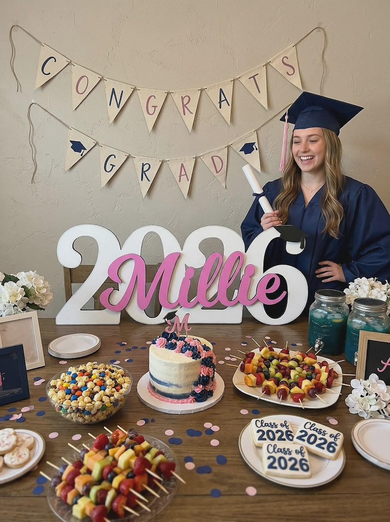 May include: A graduation celebration featuring a banner that reads "CONGRATS GRAD". A large "2026" sign with the name "Millie" in pink is displayed. The table includes a cake, fruit skewers, cookies, and popcorn.