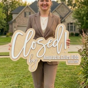 Op de afbeelding: Een licht houten bord met witte 3D-letters met de tekst Closed! by Amyracle Coles Bibb REALTOR. Het bord heeft een natuurlijke houtnerfafwerking en wordt vastgehouden door een persoon in een beige blazer en bijpassende broek.