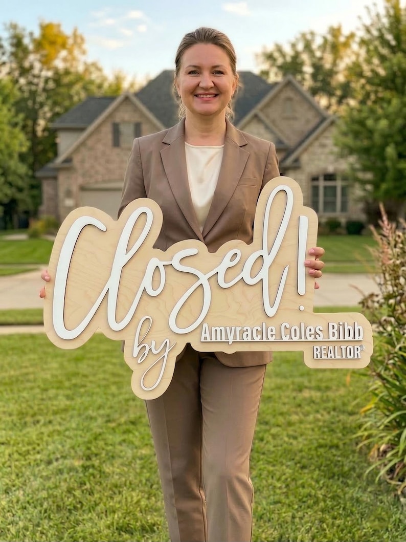 Op de afbeelding: Een licht houten bord met witte 3D-letters met de tekst Closed! by Amyracle Coles Bibb REALTOR. Het bord heeft een natuurlijke houtnerfafwerking en wordt vastgehouden door een persoon in een beige blazer en bijpassende broek.