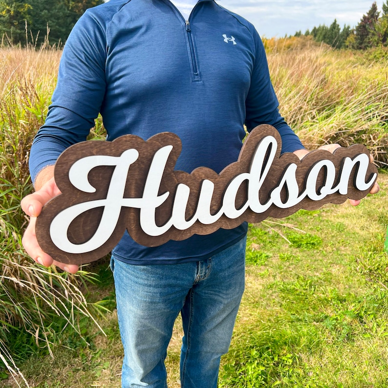 May include: A custom wooden sign featuring the name Hudson in white script lettering mounted on a dark brown stained wooden backing with a scalloped edge. The sign is held horizontally, showcasing the contrast between the white letters and wood.