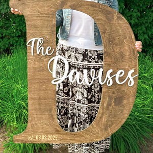 May include: A large wooden letter "D" with the words "The Davises" in white script. The date "est. 08.02.2025" is at the bottom. The wood has a natural grain and a dark brown stain. A decorative piece for a home.