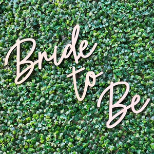 May include: Wooden sign with the words "Bride to Be" in a cursive font. The sign is light-colored and set against a backdrop of small, green leaves. The sign is likely for a bridal shower or wedding.