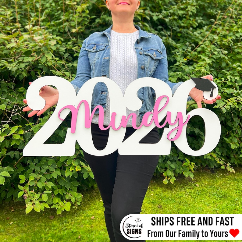 May include: A white wooden sign with the numbers "2026" and the name "Mindy" in pink. A graduation cap is attached to the number 6. The sign is held in front of a green leafy background. The sign says "SHIPS FREE AND FAST From Our Family to Yours".