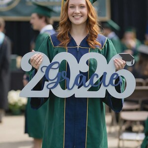 May include: A graduate in a green and navy graduation gown and cap holds a sign that says "2020 Gracie". The sign is light gray with the name in navy blue script. The graduate is smiling at the camera.