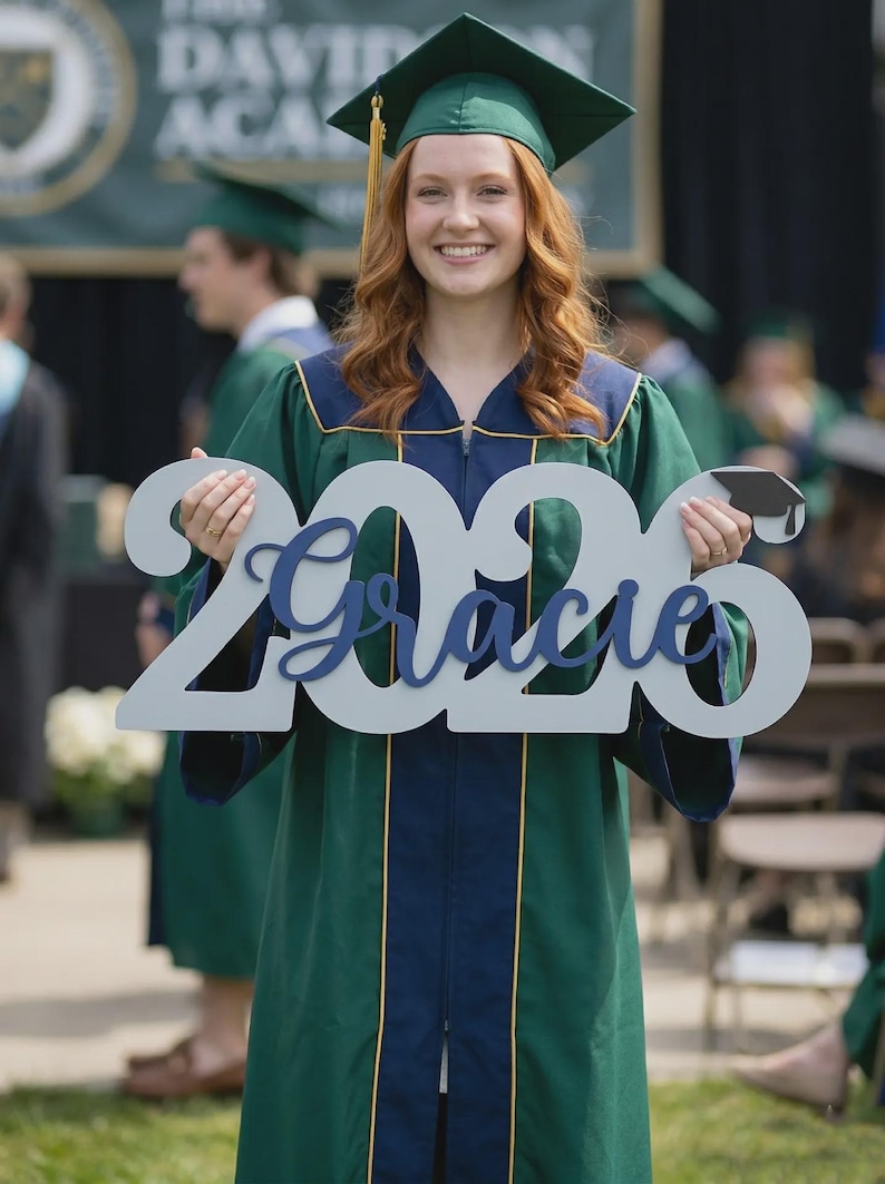 May include: A graduate in a green and navy graduation gown and cap holds a sign that says "2020 Gracie". The sign is light gray with the name in navy blue script. The graduate is smiling at the camera.