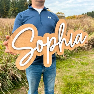 May include: A custom wooden sign featuring the name Sophia in white script lettering mounted on a light brown wooden backing with a smooth finish. The sign has a layered design with the white letters raised above the wooden base.